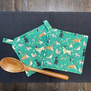 May include: Two green quilted pot holders with a pattern of various dog breeds and shamrocks. A wooden spoon rests on a black placemat next to the pot holders. The pot holders have a loop for hanging.