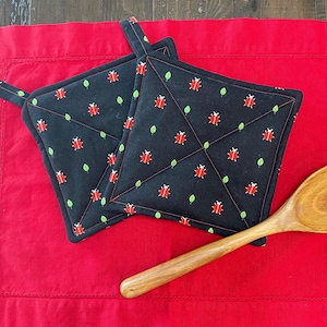 May include: Two black quilted pot holders with a red ladybug and green leaf pattern. The pot holders are on a red surface with a wooden spoon.