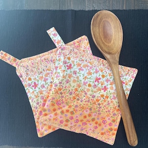 May include: Two floral print pot holders with a matching hanging loop, featuring a pattern of pink, orange, and white flowers. A wooden spoon rests beside the pot holders. The items are on a dark surface.