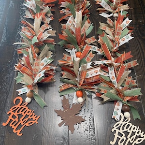 May include: Three decorative garlands made of layered fabric ribbons in shades of green, orange, and tan, featuring pumpkin and leaf patterns. Each garland includes wooden beads and a cutout shape reading Happy Fall or a maple leaf.