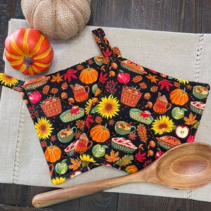 May include: Two black pot holders with a fall-themed print featuring pumpkins, leaves, pies, and sunflowers. The pot holders are on a beige linen surface with a wooden spoon.