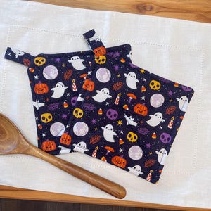 May include: Two square fabric pot holders featuring a dark purple background with a repeating pattern of white ghosts, orange pumpkins, yellow skulls, purple skulls, candy corn, stars, moons, and candles, each with a small fabric hanging loop.