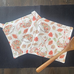 May include: Two square quilted pot holders featuring a cream-colored fabric printed with a pattern of hedgehogs, acorns, and autumn leaves in shades of orange, red, brown, and green, accompanied by a single wooden spoon placed diagonally across them.