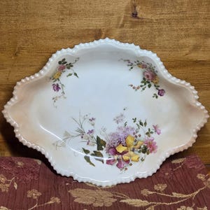 May include: An antique, white porcelain serving dish with a scalloped edge and floral design. The dish features a delicate pattern of pink, yellow, and purple flowers. The dish is approximately 12 inches in diameter.