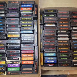 May include: A large collection of rectangular black Atari 2600 video game cartridges arranged in two cardboard boxes. Each cartridge features a label with the game title printed in various colors, including Pac-Man, Asteroids, and E.T.