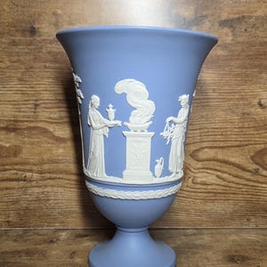 May include: A light blue vase featuring white relief figures of two people standing on either side of a central pedestal with a smoking urn. The vase has a flared rim, a pedestal base, and a decorative white band encircling the lower section.