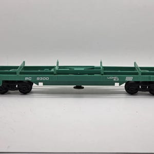 May include: A green toy train flatcar with black wheels. The flatcar has the text "PC 9300" and "LIONEL" printed on the side. The flatcar is designed to carry other toy train cars or cargo. The toy train is a model of a real-life flatcar.
