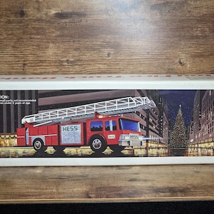 May include: A rectangular white cardboard box featuring a graphic of a red Hess fire truck with an extended ladder. The box displays the text CAUTION: Due to small parts, not recommended for children under 3 years of age and the Hess logo.