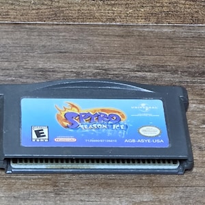 Puede incluir: Un cartucho gris de Game Boy Advance para el juego "Spyro Season of Ice". La etiqueta presenta el título del juego en morado y amarillo, con un fondo azul y blanco. El cartucho está clasificado como "Everyone" por la ESRB.