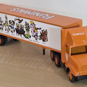 May include: Orange and white Funimals toy truck with a trailer. The trailer features cartoon animal characters and the Funimals logo. The truck has black wheels and a detailed front.