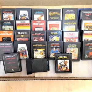 Puede incluir: Una colección de cartuchos de videojuegos Atari 2600 rectangulares y negros dispuestos en una caja de cartón. Los cartuchos presentan varias etiquetas coloridas con títulos que incluyen Air Sea Battle, Pac-Man, Space Invaders, Superman y Warlords.