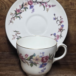 Puede incluir: Juego de taza y platillo de té blanco con diseño floral. La taza y el platillo tienen un borde dorado. El patrón floral incluye flores rosas, moradas, azules y amarillas con hojas verdes. El juego está sobre una superficie de madera.