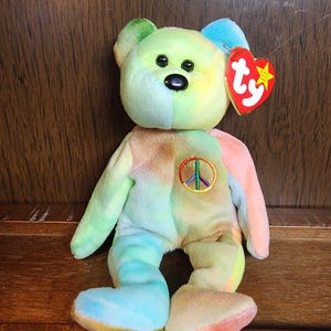 May include: A tie-dye plush bear with a peace sign on its chest. The bear is sitting on a wooden surface and has a Ty tag attached to its ear.