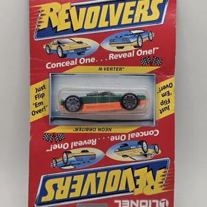 May include: A toy car on a red and yellow cardboard backing. The car is green and orange, with black wheels. The packaging reads "REVOLVERS" and "N-VERTER" with the text "Conceal One... Reveal One!"