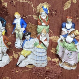 May include: A collection of ten porcelain figurines depicting people in historical attire. The figures feature various poses and colorful outfits, including blue, yellow, green, and white garments. Each figurine stands on a base, showcasing detailed painted facial features and clothing textures.