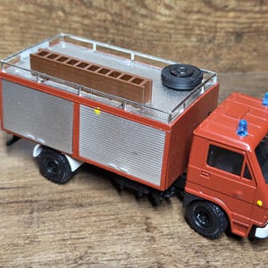 Puede incluir: Un modelo en miniatura de un camión de bomberos en rojo, plateado y marrón. El camión tiene una escalera en la parte superior, una rueda de repuesto y luces de emergencia azules. Las ruedas son negras y el camión tiene un parachoques delantero blanco.