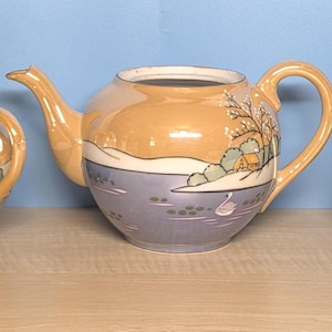 May include: A vintage tea set with a teapot, sugar bowl, and creamer. The set features a landscape design with a lake, trees, and a swan. The colors include orange, blue, and white. The set is made of ceramic.