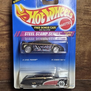 May include: A Hot Wheels Steel Stamp Series package containing two die-cast metal cars. The blue packaging displays the Hot Wheels logo and the text "FREE BONUS CAR!". The cars are black with colourful details.