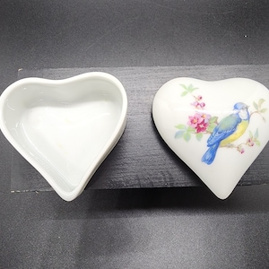 May include: White porcelain heart-shaped trinket box with a blue bird and pink flowers painted on the lid. The box is open, revealing the empty interior.