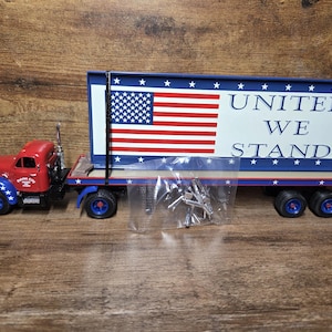 May include: A red and blue toy truck with an American flag design. The trailer features the flag and the words "UNITED WE STAND". The truck has blue wheels and a red cab with blue star accents.