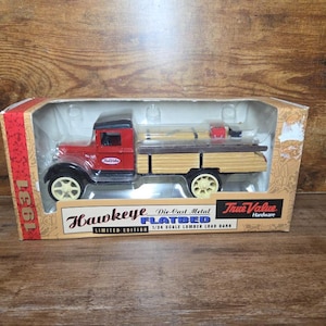 Puede incluir: Un camión Hawkeye Flatbed de metal fundido a presión, rojo con guardabarros negros y ruedas color crema, en su embalaje original. El camión tiene una carga de madera y la caja dice "1931" y "Edición Limitada".
