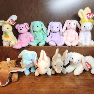 May include: A collection of ten plush bunny toys in various pastel colors, including pink, blue, green, and white. The bunnies have embroidered faces and are holding small red heart-shaped tags.