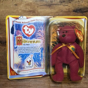 Puede incluir: Un oso de peluche Millennium Bear Ty Beanie Baby de color burdeos en su embalaje original. El embalaje incluye texto y gráficos, incluido el logotipo de McDonald's y la frase "Ronald McDonald House Charities."