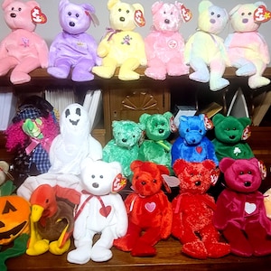 Puede incluir: Una colección de juguetes de peluche coloridos, incluyendo osos de peluche en varios colores como rosa, morado y amarillo, y otras figuras como un fantasma y una calabaza. Los juguetes están colocados sobre una superficie de madera.