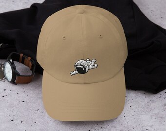 May include: A tan dad hat with an embroidered image of an astronaut floating in space. The astronaut is wearing a white spacesuit with a black visor.