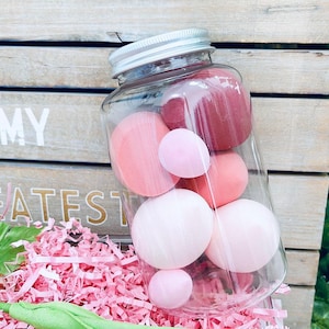 Op de afbeelding: Een glazen pot met een zilveren deksel gevuld met roze en rode zachte ballen. De pot staat op een roze en witte gescheurde papieroppervlak.