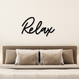 May include: A bedroom with a white wall and a bed with a wooden frame. The bed has a white duvet and brown pillows. There are two black pendant lights hanging from the ceiling. The word "Relax" is written in black script on the wall above the bed.