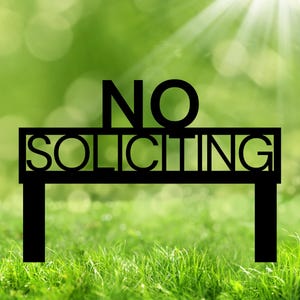 May include: A black metal sign with the words "NO SOLICITING" cut out. The sign has a rectangular shape with two vertical posts. The background is a blurred green with sunlight.