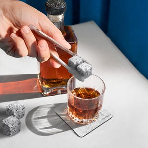May include: A set of gray stone whiskey chilling cubes being placed into a glass of amber colored whiskey with a pair of white tongs. The whiskey is in a clear glass on a gray coaster with the word "remarkable" printed on it.