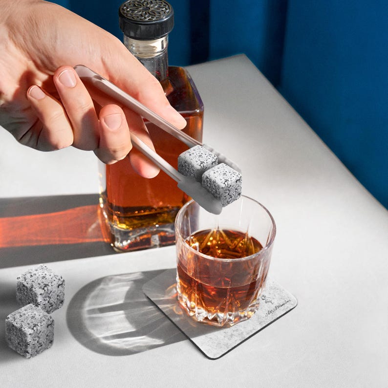 May include: A set of gray stone whiskey chilling cubes being placed into a glass of amber colored whiskey with a pair of white tongs. The whiskey is in a clear glass on a gray coaster with the word "remarkable" printed on it.