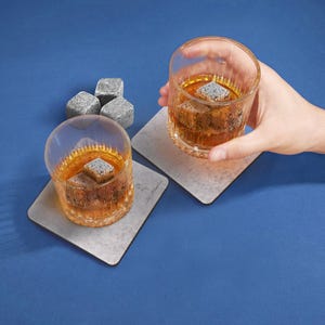 May include: Two glasses of whiskey with whiskey stones on coasters. The coasters are gray and have a speckled pattern. The glasses are clear and have a ribbed design.