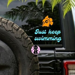 May include: Black Jeep SUV with a spare tire. A window decal features an orange fish and the words "Just keep swimming" in turquoise. A small pink sticker with a black silhouette is also visible.