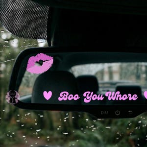 May include: A car's rearview mirror with the text "Boo You Whore" in pink script, flanked by pink hearts. A pink kiss graphic and a small sticker are also visible. The background shows a rainy, blurred outdoor scene.