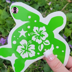 May include: A green turtle-shaped sticker with white hibiscus flowers, a starfish, and polka dots. The sticker is held by a hand with a white and pink manicure. The background is blurred green foliage.