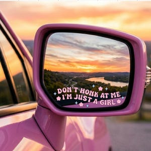 May include: Pink car side mirror with the text "DON'T HONK AT ME I'M JUST A GIRL" in a whimsical font. The mirror reflects a scenic sunset over a river and hills.