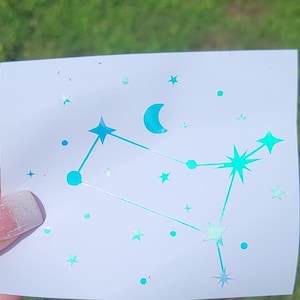 May include: A square sticker featuring a teal constellation design with stars, dots, and a crescent moon. The sticker has a holographic, iridescent finish, creating a shimmering effect. The design is set against a white background.
