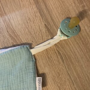 May include: A light blue and white muslin baby blanket with a pacifier attached. The blanket has a white tag with the text "Handmade with &hearts; by Graine de Bonheur".