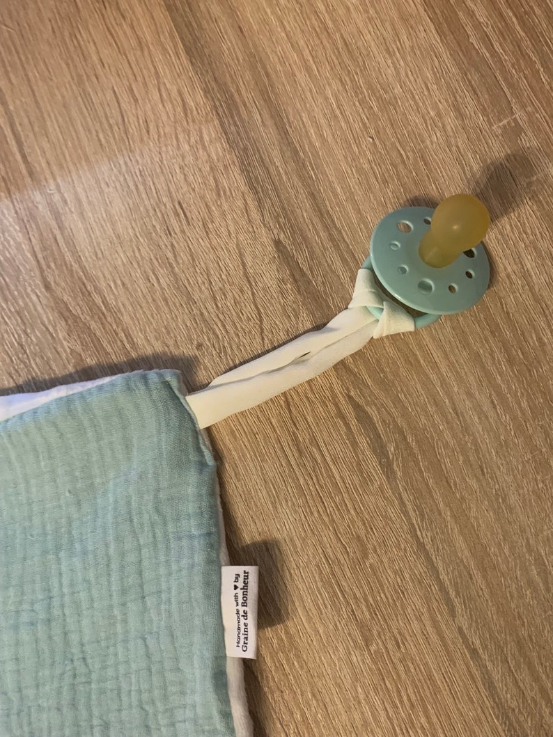 May include: A light blue and white muslin baby blanket with a pacifier attached. The blanket has a white tag with the text "Handmade with &hearts; by Graine de Bonheur".