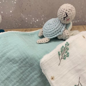 May include: A light blue crocheted turtle toy on a light blue and white baby blanket with a floral pattern.
