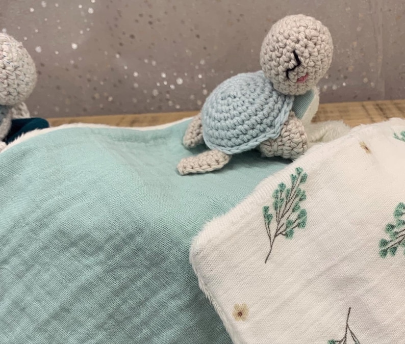 May include: A light blue crocheted turtle toy on a light blue and white baby blanket with a floral pattern.