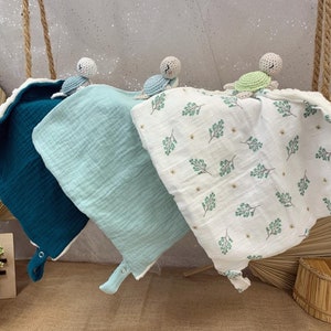 May include: Three crocheted turtle toys with different colored shells, teal, light blue, and green, are attached to three muslin baby blankets. The blankets are layered on top of each other and have different patterns. The top blanket is white with a green leaf pattern, the middle blanket is light blue, and the bottom blanket is teal.