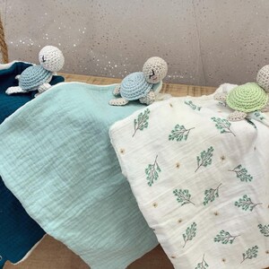 May include: Three crocheted turtle toys with different colored shells, green, white, and gray, are placed on top of three different colored baby blankets. The blankets are teal, light blue, and white with a green leaf pattern.