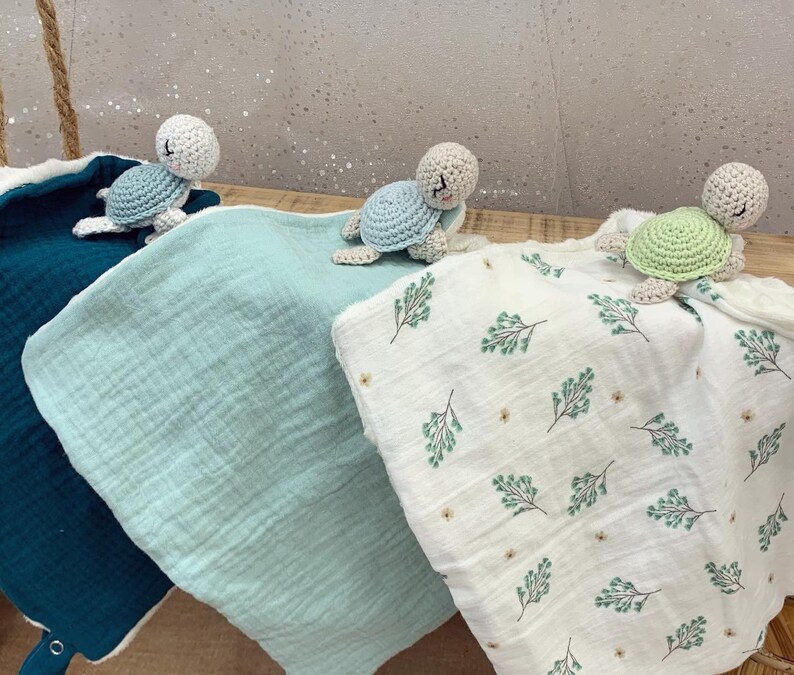 May include: Three crocheted turtle toys with different colored shells, green, white, and gray, are placed on top of three different colored baby blankets. The blankets are teal, light blue, and white with a green leaf pattern.
