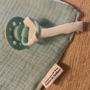 May include: A light blue muslin baby blanket with a white border and a pacifier clip attached. The blanket has a label that reads "Handmade with &hearts; by Graine de Bonheur".