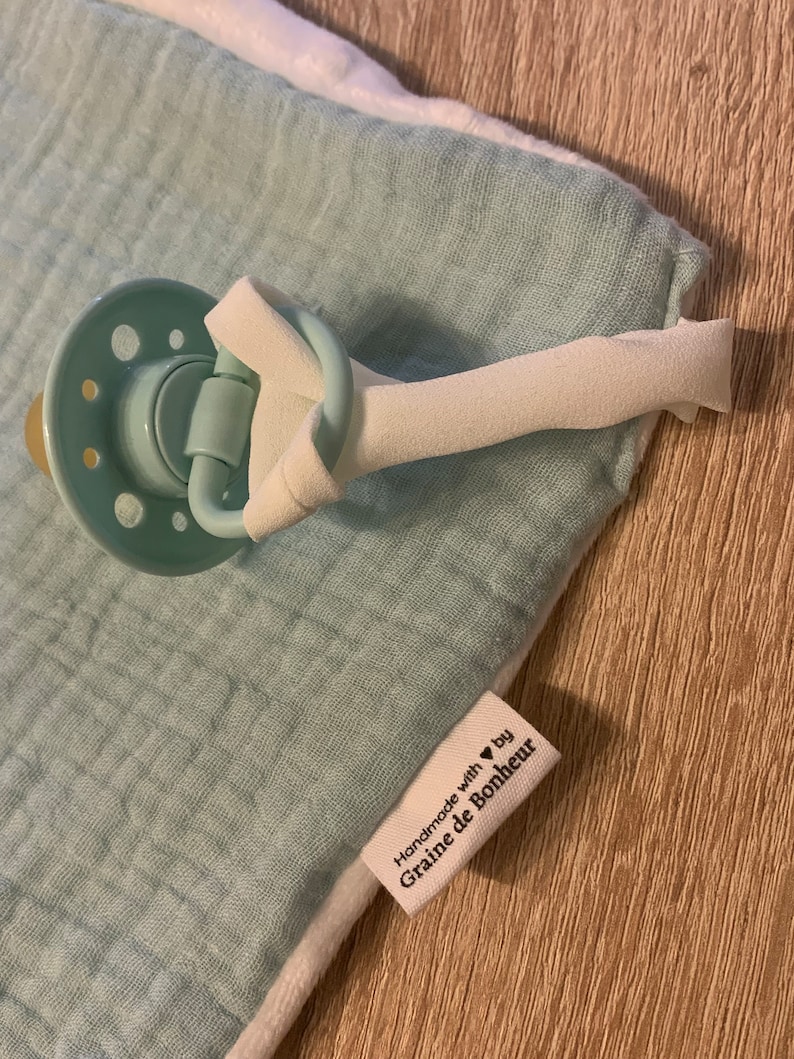 May include: A light blue muslin baby blanket with a white border and a pacifier clip attached. The blanket has a label that reads "Handmade with &hearts; by Graine de Bonheur".