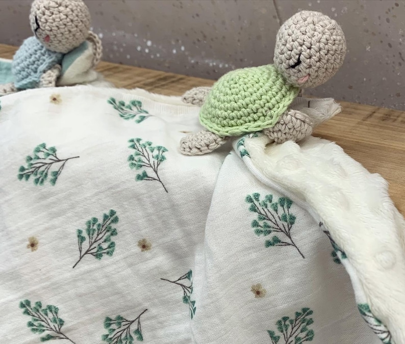 May include: A white baby blanket with a green leafy pattern and two crocheted turtle toys. One turtle is green and the other is beige.